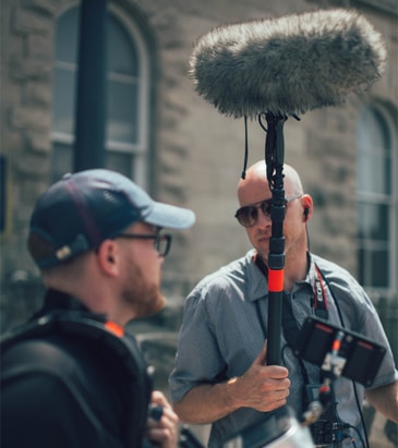 A man holding a Boom pole with a shotgun-mic attached on to it via a pistol grip mount. The mic is probably on a shockmount but is covered in a furry wind muff. The mic is connected to a field recorder via XLR cable.